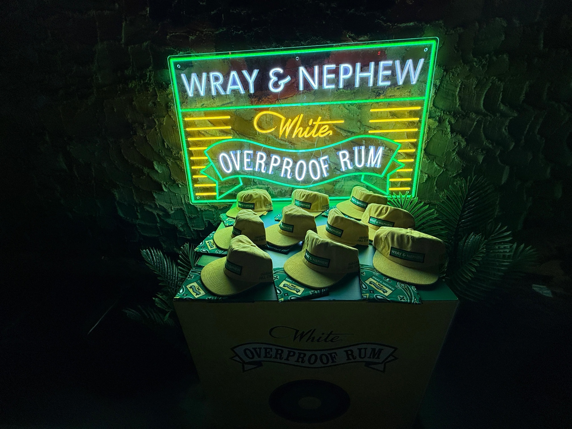 Branded yellow corduroy hats displayed on a custom crate beneath a Wray and Nephew neon sign
